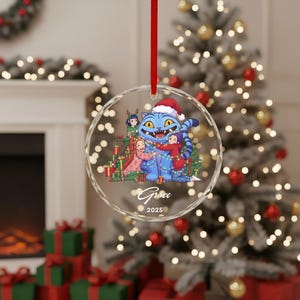 May include: A clear, round Christmas ornament with a red ribbon. The ornament features a blue cat wearing a Santa hat, surrounded by children and Christmas presents. The name "Grace" and the year "2025" are written on the ornament.