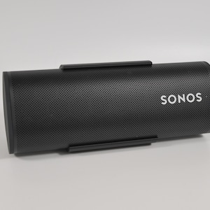 May include: A black, cylindrical speaker with the brand name "SONOS" in white lettering. The speaker has a perforated surface and two black handles. The speaker is designed to be portable.