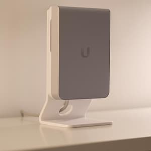 Puede incluir: Un dispositivo de red blanco y gris con una base curva y una parte superior rectangular. El dispositivo tiene una parte frontal gris con un pequeño logotipo "U". Está sobre una superficie blanca.