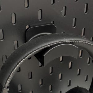 Może przedstawiać: Czarne słuchawki nauszne wiszące na czarnej tablicy perforowanej. Słuchawki są zamontowane na czarnym uchwycie. Tablica perforowana ma równomiernie rozmieszczone, podłużne otwory. Poniżej widoczna jest biała powierzchnia.