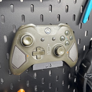 Puede incluir: Un mando Xbox verde oliva está montado en una placa perforada negra. El mando tiene detalles grises y un diseño de flecha blanca. La placa perforada tiene agujeros espaciados uniformemente y se utiliza para organizar objetos.