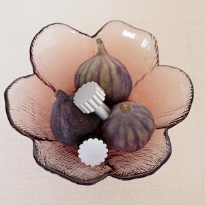 Pode incluir: Uma tigela de vidro rosa em forma de flor contém vários figos maduros e dois objetos decorativos prateados. Os figos são roxos escuros com listras verdes. A tigela está sobre uma superfície bege clara.