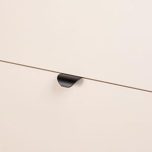 Op de afbeelding: Een close-up van een moderne kastdeur met een matte, gebroken witte afwerking. Een slanke, zwarte, gebogen handgreep is horizontaal op de deur gemonteerd. Het ontwerp van de handgreep is minimalistisch en eigentijds.