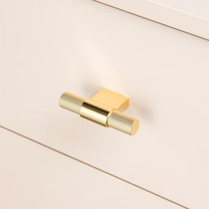 Può includere: Una maniglia per mobili color oro con un design a T. L'impugnatura cilindrica presenta una texture a coste e la base è un blocco rettangolare. La maniglia è montata su una superficie di colore chiaro.
