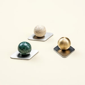 Puede incluir: Tres esferas decorativas: una bola de mármol verde, una piedra beige y una bola de metal dorado. Cada esfera descansa sobre una pequeña base cuadrada de metal cepillado. Fondo crema.