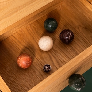 Op de afbeelding: Een houten lade gevuld met zes decoratieve stenen bollen in verschillende maten en kleuren. De bollen hebben groene, witte, bruine en oranje tinten. De lade is gemaakt van lichtgekleurd hout.