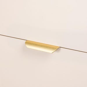 May include: A gold-colored cabinet pull with a curved design. The pull is mounted on a light-colored cabinet door. The hardware has a brushed metal finish and a modern aesthetic.