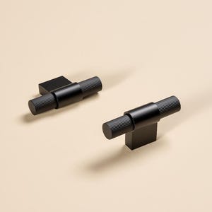 Pode incluir: Duas puxadores de armário pretos foscos com uma alça cilíndrica canelada e uma base retangular. O hardware foi projetado para fácil instalação e adiciona um toque moderno a qualquer armário ou gaveta. Os puxadores são mostrados em um fundo neutro.