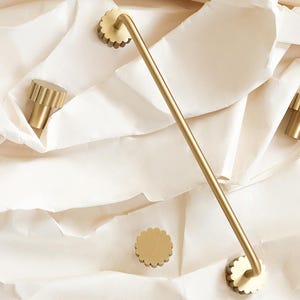 Può includere: Una collezione di accessori per il bagno color oro, tra cui un portasciugamani e pomelli per armadi. Il portasciugamani è dritto con estremità smerlate. I pomelli hanno un design a coste. Gli articoli sono disposti su una superficie strutturata color crema.