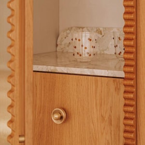 Op de afbeelding: Een houten kast met een lade en een ronde knop. Twee heldere glazen met oranje stippenpatronen staan op een marmeren plank. De kast heeft een decoratieve, golvende rand.