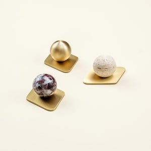 Könnte beinhalten: Drei dekorative Objekte. Jedes hat eine Kugel auf einer quadratischen, goldfarbenen Basis. Die Kugeln variieren: eine ist goldfarben, eine ist weiß mit braunen Sprenkeln und eine ist weinrot und weiß. Die Objekte sind auf einer cremefarbenen Oberfläche angeordnet.