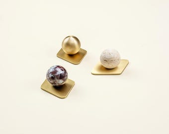 Op de afbeelding: Drie decoratieve objecten. Elk heeft een bol op een vierkante, goudkleurige basis. De bollen variëren: één is goudkleurig, één is wit met bruine spikkels en één is bordeauxrood en wit. De objecten zijn geplaatst op een crèmekleurig oppervlak.