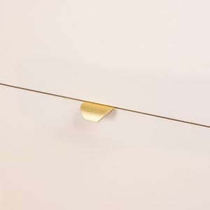 Könnte beinhalten: Nahaufnahme einer Schranktür mit einem goldfarbenen, halbmondförmigen Griff. Die Tür hat eine hellcremefarbene Farbe, und der Griff befindet sich auf der rechten Seite. Die Schranktür hat ein klares, modernes Design.