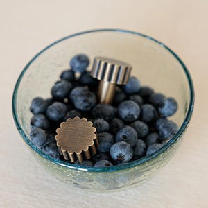 Pode incluir: Uma tigela de vidro transparente com borda azul-petróleo cheia de mirtilos frescos. Dois objetos decorativos de metal, um com a parte superior em forma de engrenagem e outro com borda ondulada, repousam entre as bagas.