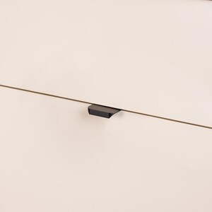 May include: Close-up of a section of a light-colored cabinet with a dark gray, minimalist handle. The handle is a small, rectangular shape, and is mounted horizontally. The cabinet appears to be made of wood or a similar material.