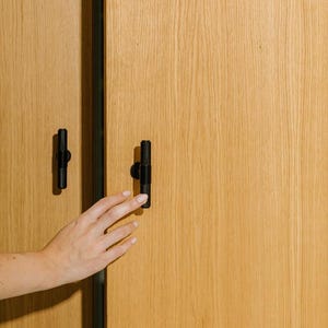 Op de afbeelding: Houten kastdeuren met zwarte hardware. De deuren zijn gemaakt van lichtgekleurd hout met een zichtbare nerf. Een hand reikt naar een van de zwarte handgrepen. De kast heeft een verticale zwarte afwerking.
