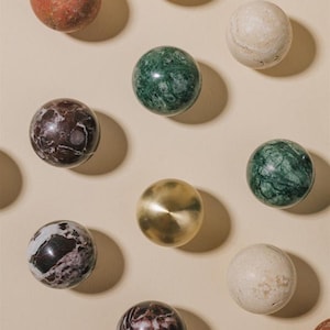 Può includere: Un assortimento di pomelli decorativi per mobili in vari materiali e colori. I pomelli includono ottone lucido, marmo con venature marroni e bianche, marmo verde e pietra bianco sporco. I pomelli sono disposti su uno sfondo neutro.