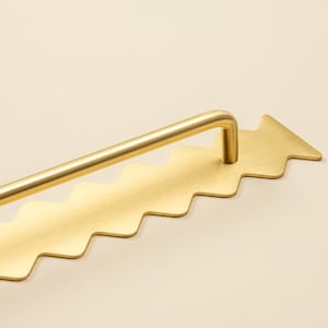 Può includere: Una maniglia per mobili in metallo color oro dal design unico. La maniglia presenta una maniglia a barra dritta e una base decorativa con un bordo a zig-zag e un'estremità a forma di freccia. L'hardware è in ottone.