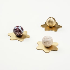 Può includere: Tre oggetti decorativi. Ognuno presenta una sfera su una base dorata a forma di stella. Le sfere sono realizzate con materiali diversi: una pietra fantasia, un metallo dorato pieno e una pietra strutturata. Sono disposti su una superficie bianca.