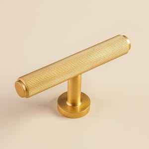 Könnte beinhalten: Ein goldfarbener Schrankgriff mit zylindrischem Griff mit geriffelter Textur. Der Griff ist an einer kreisförmigen Basis mit einem kurzen, vertikalen Pfosten befestigt. Geeignet für Schubladen und Schränke.