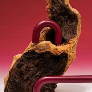 May include: A still life composition featuring a piece of weathered wood with a deep red, curved metal object. The wood has a rough, textured surface, and the background is a gradient of red and white.