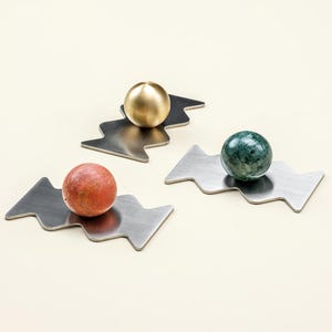 Könnte beinhalten: Drei dekorative Objekte. Jedes hat eine Metallbasis mit Zickzack-Design, die eine Kugel hält. Eine Kugel ist goldfarben, eine andere rot und die dritte grün. Die Objekte befinden sich auf einer cremefarbenen Oberfläche.