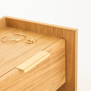 Op de afbeelding: Close-up van een houten nachtkastje met een goudkleurige ladeknop. Een gouden armband en twee ringen liggen op het oppervlak van het nachtkastje. Het nachtkastje heeft een lichte houtnerf afwerking en een eenvoudig, modern design.