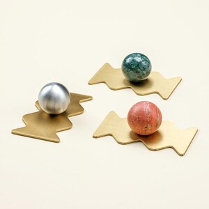 Peut inclure: Trois objets décoratifs. Chacun présente une base métallique dorée en forme de zigzag. L'une des bases contient une sphère argentée, une autre une sphère tachetée verte et noire, et la troisième une sphère rouge-orangé. Les objets sont sur une surface crème.