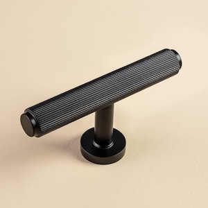 Könnte beinhalten: Ein schwarzer Schrankgriff mit einem zylindrischen, geriffelten Griff und einem kreisförmigen Sockel. Die Hardware ist aus Metall und hat eine matte Oberfläche. Der Griff ist ca. 15 cm lang.