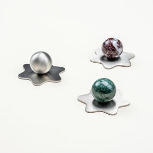 Peut inclure: Trois objets décoratifs. L'un est une sphère argentée sur une base métallique en forme d'étoile. Les deux autres sont des sphères en pierre, une rouge et blanche, l'autre verte, chacune sur une base métallique similaire. Les objets sont sur fond blanc.