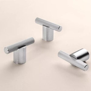 Könnte beinhalten: Drei silberfarbene Schrankknöpfe mit T-Bar-Design. Jeder Knopf hat einen zylindrischen Sockel und einen horizontalen Balken mit feinen, parallelen Rillen. Die Knöpfe sind auf einer hellbeigen Oberfläche angeordnet.