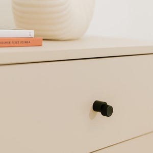 May include: A close-up of a cream-colored dresser drawer with a black cylindrical knob. A white, textured vase and two books are on the surface above the drawer. The book on the bottom has an orange cover.