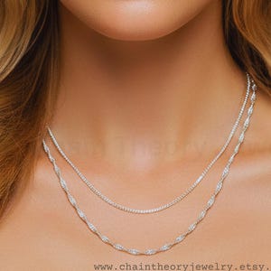 May include: Two silver necklaces with sparkling details. The top necklace is a delicate chain, while the lower one features an intricate, twisted design. The necklaces are displayed against a blurred background. The website address www.chaintheoryjewelry.etsy.com is visible at the bottom.