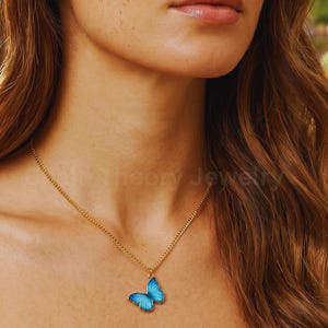 May include: A gold-toned chain necklace featuring a small, vibrant blue butterfly pendant with delicate wing details. The pendant hangs centrally from the thin metal chain, which rests against the collarbone area. The text chaintheoryjewelry.etsy.com is visible.