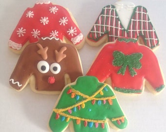 May include: Five Christmas-themed sugar cookies decorated with icing to look like ugly sweaters. One cookie has a reindeer face, one has a plaid pattern, one has a bow, one has snowflakes, and one has Christmas lights.