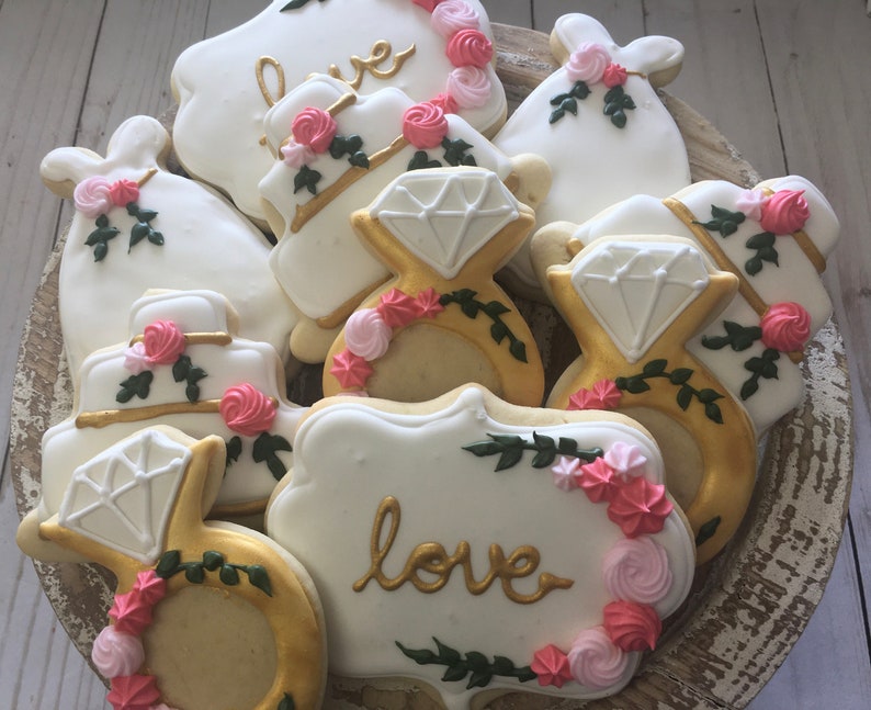 May include: A plate of decorated sugar cookies with a wedding theme. The cookies are decorated with white icing, gold accents, and pink and green details. The cookies feature images of wedding rings, cakes, and the word "love".