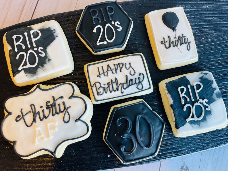 May include: A collection of seven decorated sugar cookies featuring black and white icing. Designs include text reading RIP 20's, thirty, HAPPY Birthday, thirty AF, and the number 30. The cookies are shaped as squares, hexagons, and scalloped rectangles.
