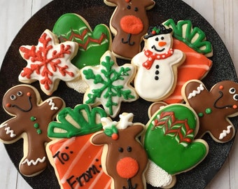 May include: A collection of Christmas-themed sugar cookies decorated with various designs, including a snowman, reindeer, snowflakes, mittens, and a gift box. The cookies are arranged on a black plate with a white background.
