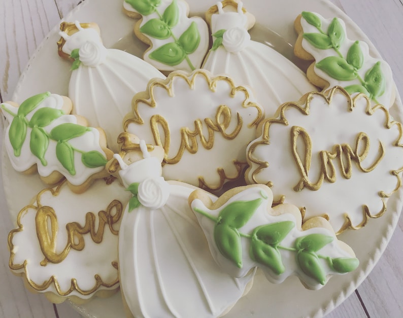May include: A plate of white and gold decorated sugar cookies with various designs, including a dress, leaves, and the word "love".