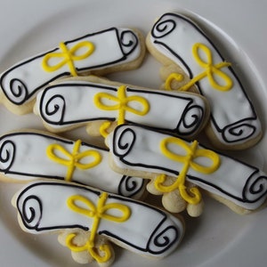 Op de afbeelding: Een bord met afstudeerkoekjes die zijn versierd met witte glazuur en gele lintjes. De koekjes zijn gevormd als opgerolde diploma's.