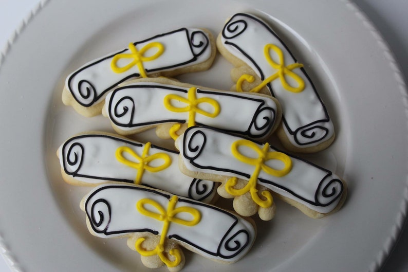 Op de afbeelding: Een bord met afstudeerkoekjes die zijn versierd met witte glazuur en gele lintjes. De koekjes zijn gevormd als opgerolde diploma's.