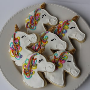 May include: Five unicorn-shaped sugar cookies decorated with white frosting and colorful swirls. Each cookie has a gold glitter horn.