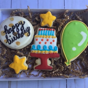 May include: A white box filled with decorated sugar cookies. The cookies include a round cookie with "happy birthday" written on it, a three-tiered cake, two yellow stars, and a green balloon. The cookies are surrounded by brown crinkle paper.