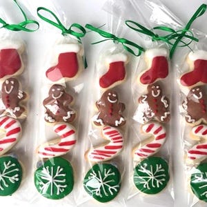 May include: Five clear plastic bags each containing four holiday-themed sugar cookies: a red stocking, a brown gingerbread person, a red and white candy cane, and a green snowflake cookie. Each bag is tied at the top with a thin green ribbon.