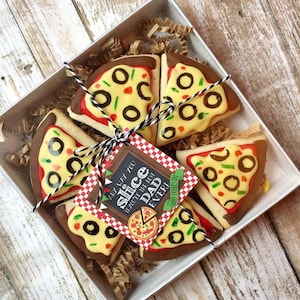 May include: A clear box filled with six decorated sugar cookies shaped like pizza slices. The cookies are decorated with yellow, brown, green, and black icing. A gift tag with the text "You are my slice of the best dad ever!" is attached to the box.