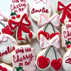 May include: Assortment of decorated sugar cookies with a cherry theme. Some cookies feature red bows, while others have cherry designs and the text "She's the Cherry on top" or the name "Amelia" in red icing. The cookies are on a white surface.