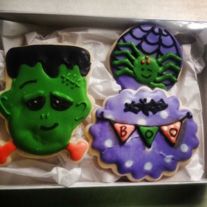 May include: A set of three Halloween-themed sugar cookies. One cookie is decorated with a green Frankenstein face, another with a purple spiderweb and a spider, and the third with a purple cookie with white polka dots and a banner that says "BOO".