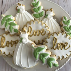May include: A plate of decorated sugar cookies with white icing and gold lettering. The cookies are shaped like leaves, dresses, and hearts. The hearts have the word "love" written on them in gold.