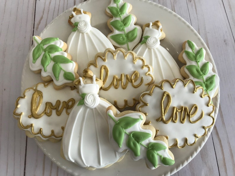 May include: A plate of decorated sugar cookies with white icing and gold lettering. The cookies are shaped like leaves, dresses, and hearts. The hearts have the word "love" written on them in gold.