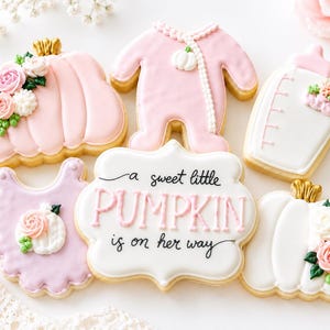May include: A collection of six decorated sugar cookies featuring a pink pumpkin with flowers, a pink baby onesie, a white baby bottle, a light purple bib with a small pumpkin, a white plaque with the text A sweet little PUMPKIN is on her way, and a white pumpkin.